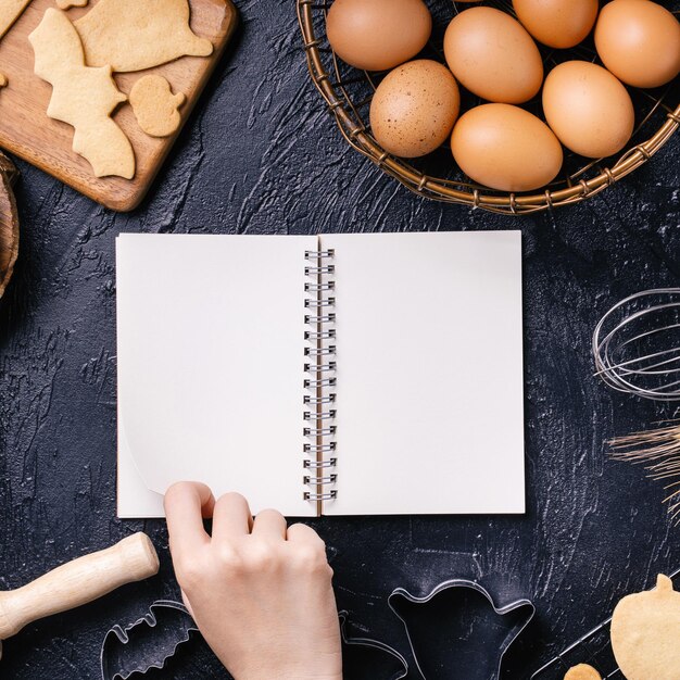 Woman is reading cookbook recipe of making Halloween cookies with baking ingredients design concept of cooking class top view flat lay overhead