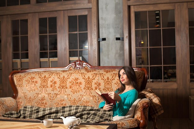 Woman is reading a red book on the beautiful sofa with   squared plaid near the table with a cup and teapot