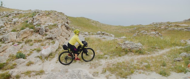 女性は、絵のように美しい谷を背景に山の湖がある丘の上に乗っています。女の子の旅行者は、バイクパッキングバッグを持った砂利ツーリングバイクで旅をします。