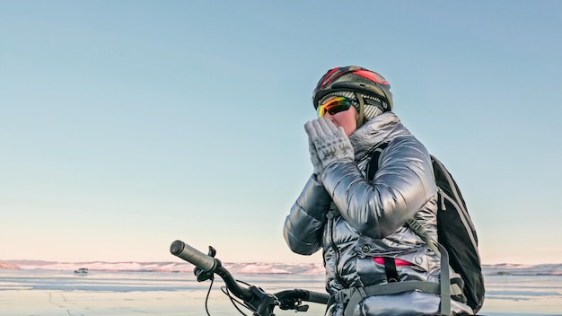 写真 女性は氷の上で自転車に乗っています女の子は銀色のダウンジャケットサイクリングバックパックとヘルメットに身を包んでいます自転車の凍ったバイカル湖のタイヤはスパイクで覆われています旅行者は乗車サイクルです