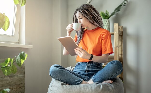 Woman is sitting on a bag chair with a cup of coffee at home\
and using a tablet