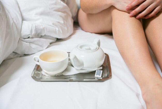 Woman is sitting in the bed. Beautiful slim legs. Girl with tea and cup on the tray. Morning at the hotel.