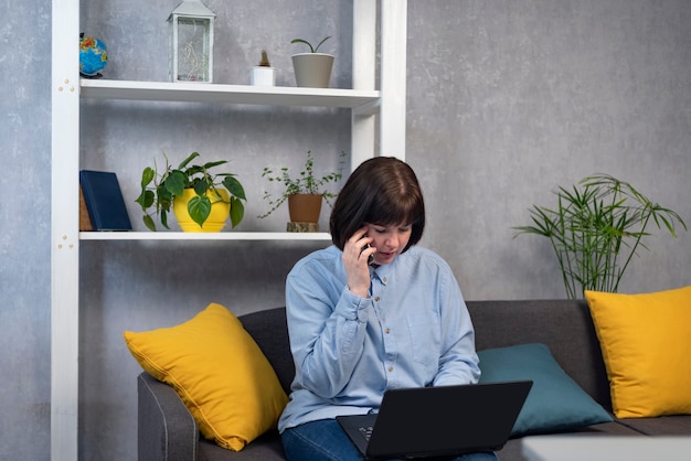 Woman is sitting on couch with laptop and talking on phone. Business woman freelancer. Entrepreneur solves work issues