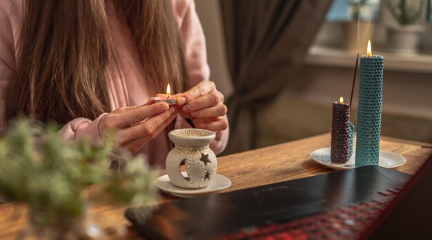 Woman is sitting in front of a laptop lighting a candle for an aroma lamp before an instructor's video lecture and creating an atmosphere for meditation and relaxation