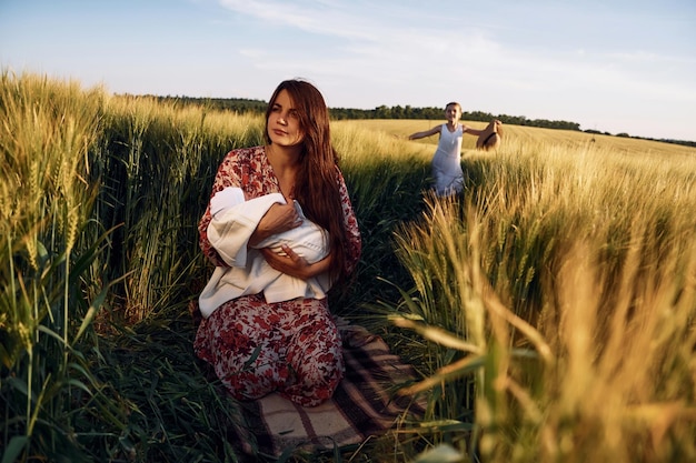 Woman is sitting on the ground Mother with her newborn baby and girl is on the field at sunny day together