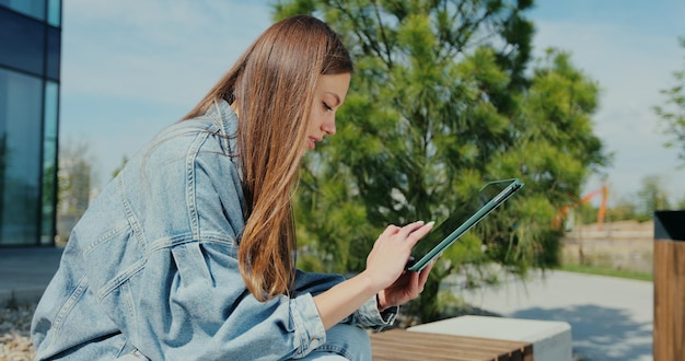 Woman is sitting on a park bench and scrolling on the tablet in her hands Attractive female messaging and chatting on tablet computer in city