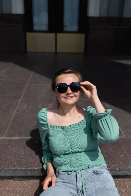 A woman is sitting on the steps near the theater and wearing sunglasses