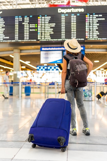 A woman is standing in an airport with a blue suitcase and a hat on