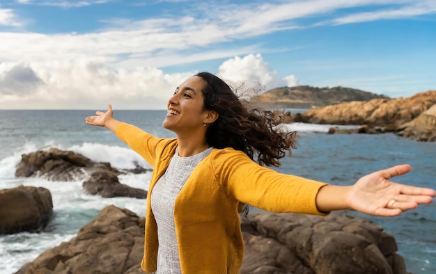 A woman is standing on a rocky beach smiling and waving her arms in the air