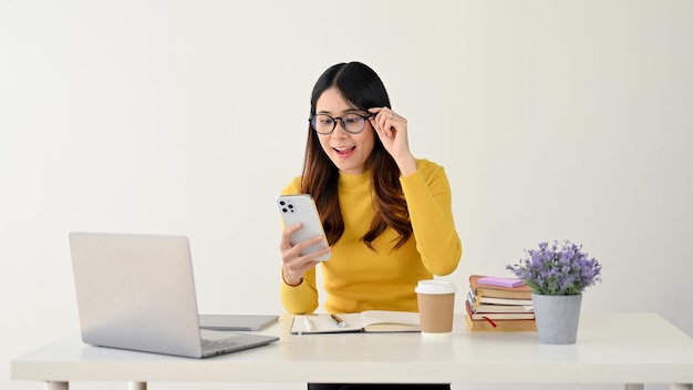 A woman is touching her glasses while looking at her phone screen reading messages