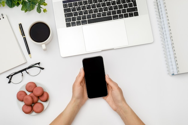 La donna sta usando lo smartphone con lo schermo in bianco sopra il tavolo bianco della scrivania con il computer portatile