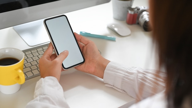 A woman is using a white blank screen smartphone while sitting in front of a computer monitor