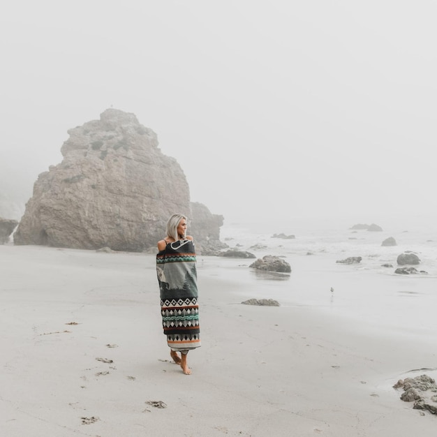 a woman is walking on the beach in front of a rock.