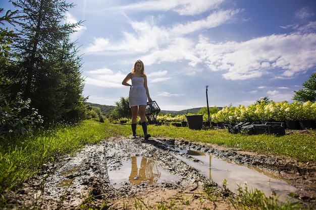 The woman is wearing white dress and rubber boots walks at the mud The gardener holds a plastic box and walks near the plantings