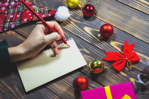 Woman is writing Christmas postcards. Greetings with holidays. Happy new year time. Congratulations to friends and family. Decorations on wooden table. Cozy mood. Copy space place.