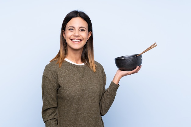 Woman over isolated blue wall smiling a lot while holding a bowl of noodles with chopsticks