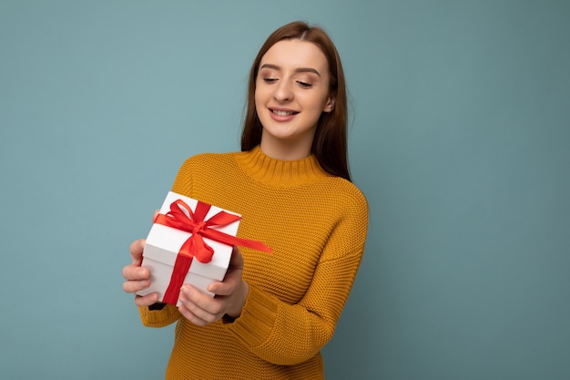 Donna isolata su un muro colorato che indossa abiti alla moda guardare tenendo la confezione regalo e guardando la scatola attuale con il nastro rosso.