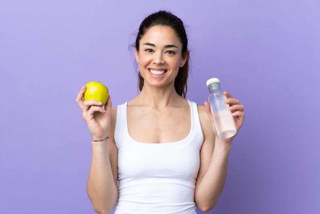 Woman over isolated purple with an apple and with a bottle of water