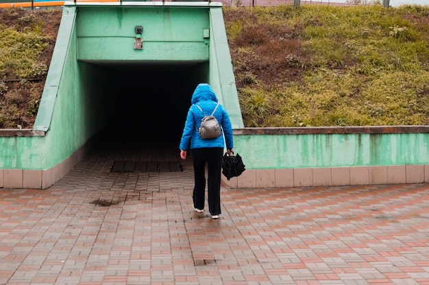 비가 오는 동안 재킷을 입은 여성이 도시의 도시에서 우산을 들고 길 아래 터널로 들어갑니다.