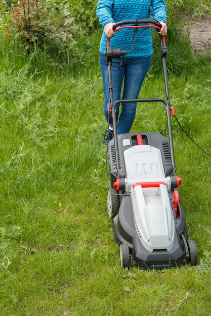 Woman in jeans is operating with lawn mower in the garden in sunny summer day. Mower grass equipment. Mowing gardener care work tool.