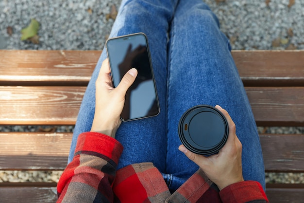 Woman in jeans and shirt hold paper cup and phone outdoor