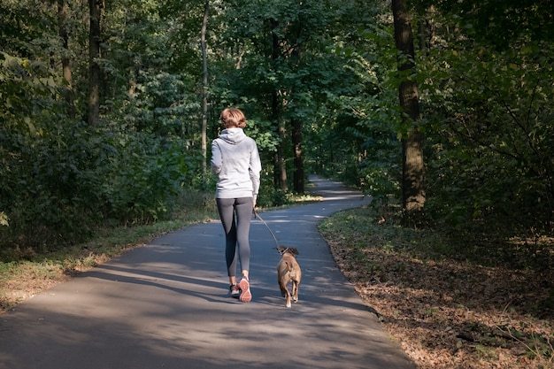 美しい森で犬とジョギングの女性。新鮮な空気で運動を実行しているクロスカントリーをやっているペットを持つ若い女性人。