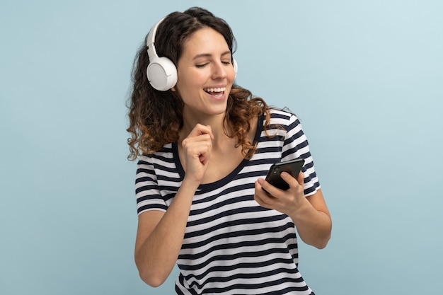 Woman joyful wearing wireless headphones listening to music, holding mobile phone in hand, dancing