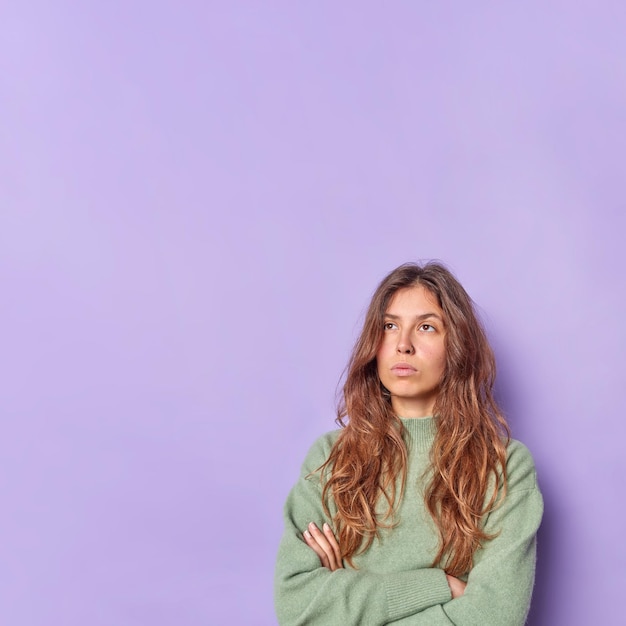 Woman keeps arms folded concentrated above thinks about something wears casual jumper isolated on purple with copy space for your advertising content