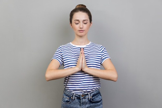 Woman keeps hands in yoga gesture has calm facial expression\
keeping palms pressed together