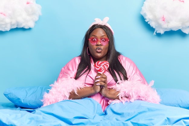 woman keeps lips folded holds delicious sweet lollipop wears heart shaped sunglasses and dressing gown poses on comfortable bed under warm blanket has lazy day