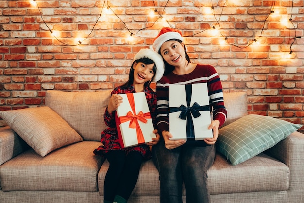 여자와 아이 배경 얼굴 카메라 미소에 붉은 벽돌 벽 실내 소파에 앉아 선물을 들고. 박싱 데이에 즐겁게 크리스마스를 축하하는 선물 상자를 보여주는 가족.
