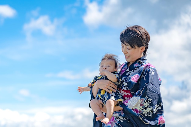 A woman in a kimono holds a baby in front of a blue sky.