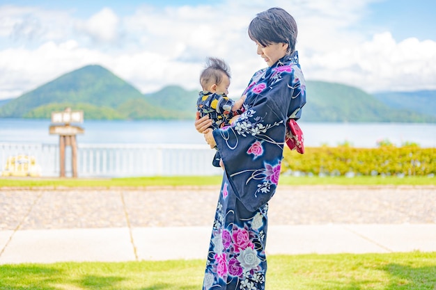 A woman in a kimono holds a baby in front of a lake.