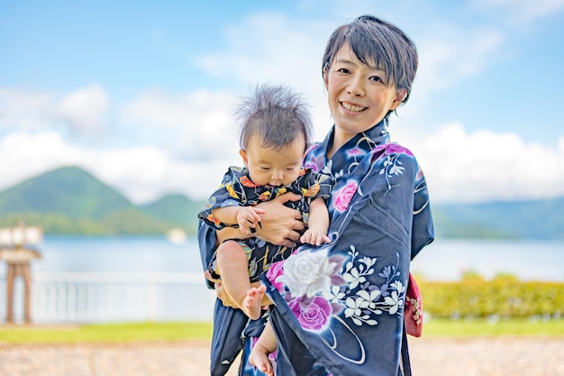 A woman in a kimono holds a baby in front of a lake.