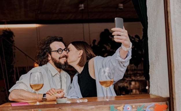Woman kissing man in a cheek and take a selfie while they drinking wine in bar during romantic date