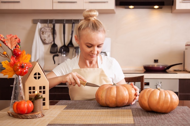 Woman on kitchen carves a pumpkin for Halloween in a room with autumn decor and a lamp house. Cosy home and preparing for Halloween.