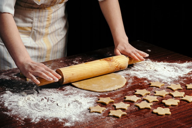 Woman in kitchen is rolling dough on wooden table with wooden rolling pin