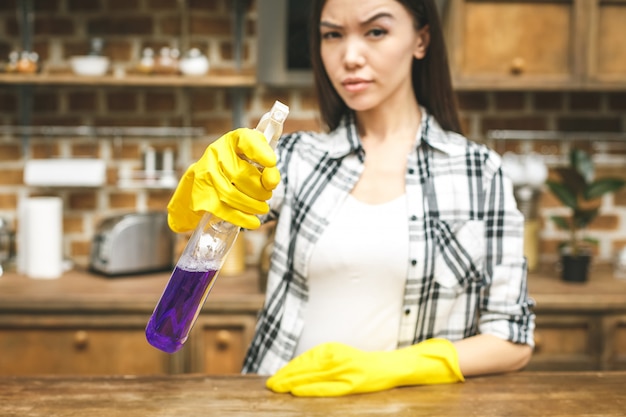 Woman in the kitchen is smiling and wiping dust using a spray and a duster while cleaning her house, close-up