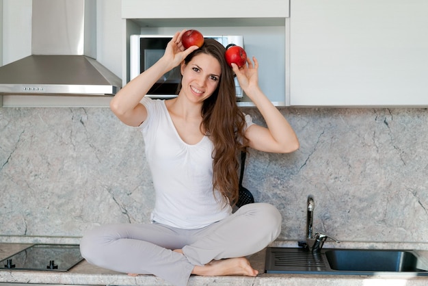 Woman in kitchen sits on table girl holds apples next to her head instead her