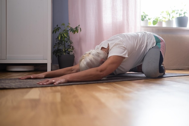 Woman kneeling on a mat and doing a balasana asana while practicing yoga at home