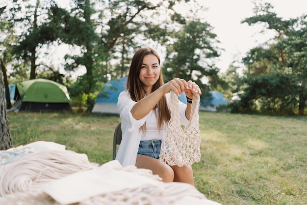 屋外のテント近くでマクラメ技術を使ってバッグを編む女性 アウトドア趣味のマクラメ編み