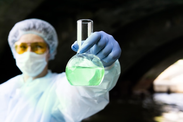 Woman in laboratory protective suit shows a flask with greenish liquid