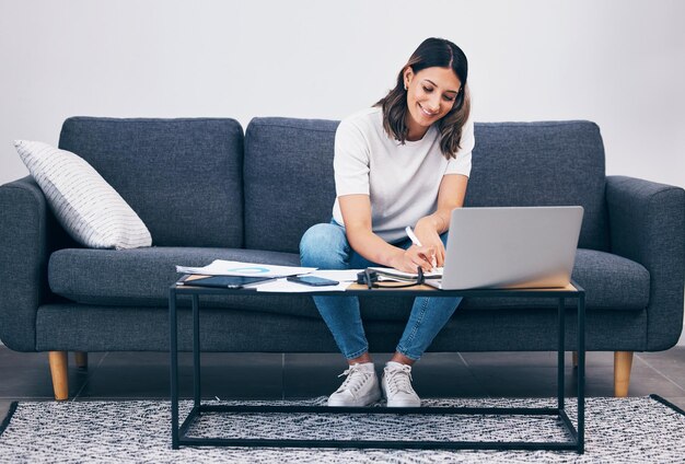 Woman laptop and writing notes on living room sofa in remote work or studying on table at home Female freelancer working with smile for planning strategy or notebook by computer sitting on couch