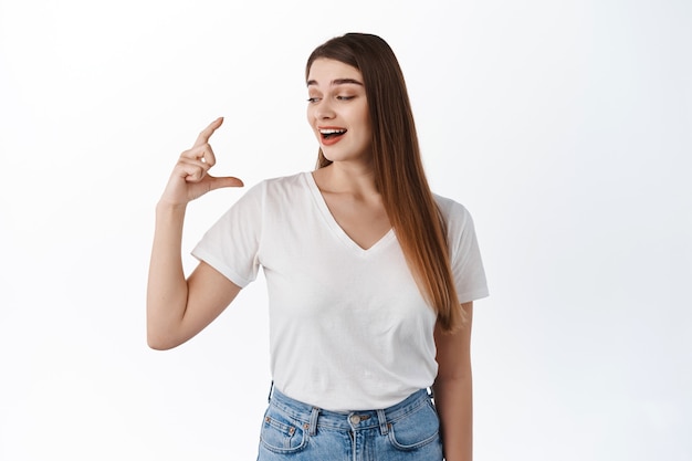 Woman laughing at small size thing, showing tiny little object in hand and looking skeptical, standing in t-shirt against white wall