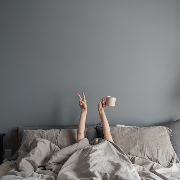 Woman laying in bed, holding mug with coffee in hand and show\
victory symbol