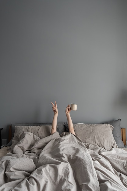 Woman laying in bed and holding mug with coffee with hand and show Victory symbol