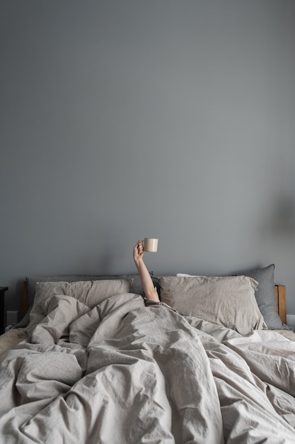 Woman laying in bed and holding mug with coffee with hand.