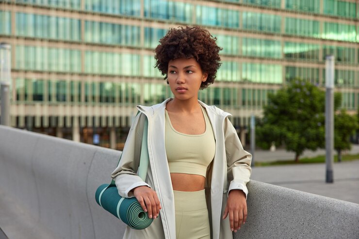 woman leads sporty lifestyle carries rolled fitness mat concentrated away with thoughtful expression dressed in sportswear poses outdoors