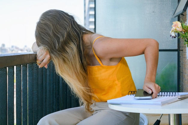 Woman leaning on the balcony railing at home tired from working so much on her phone