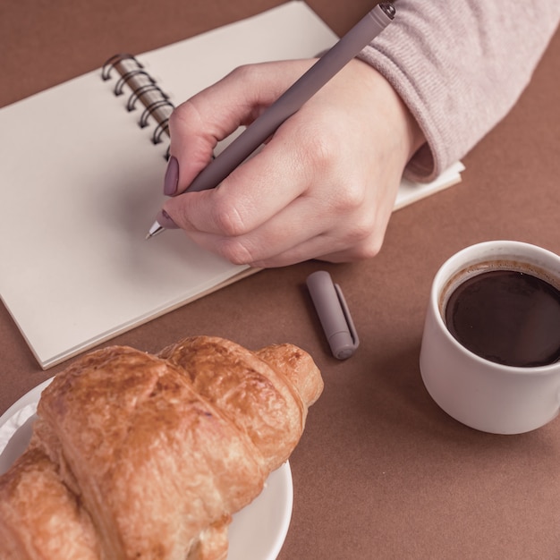 Woman left hand with pen writing on notebook at coffee shop. Freelancer working in outdoor. Coffe break with croissant and espresso